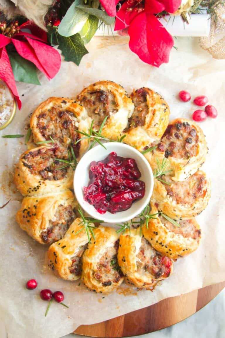 Circular sausage roll wreath on a platter, surrounded by bright cranberry dipping sauce