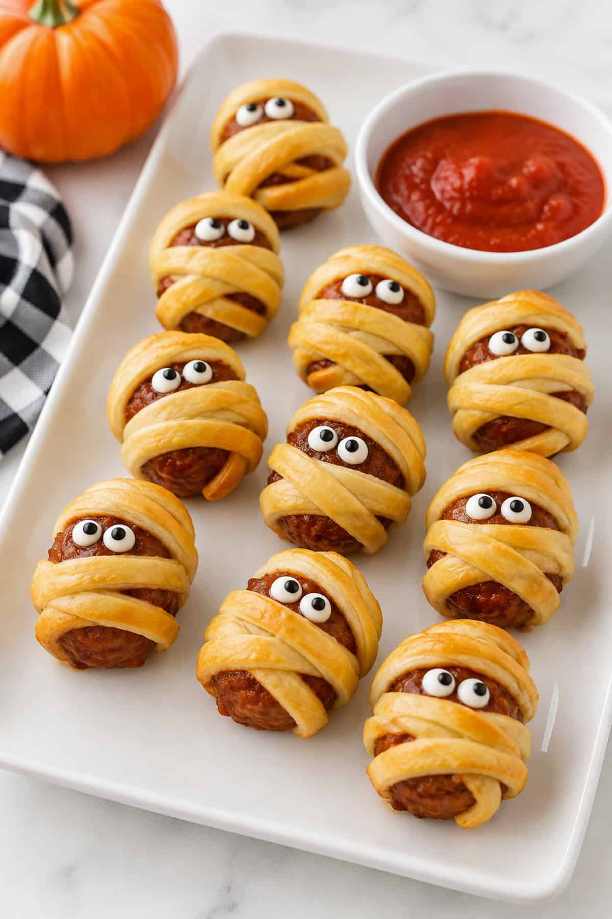 Meatballs wrapped in strips of golden crescent dough resembling mummies with red sauce eyes on a baking sheet.